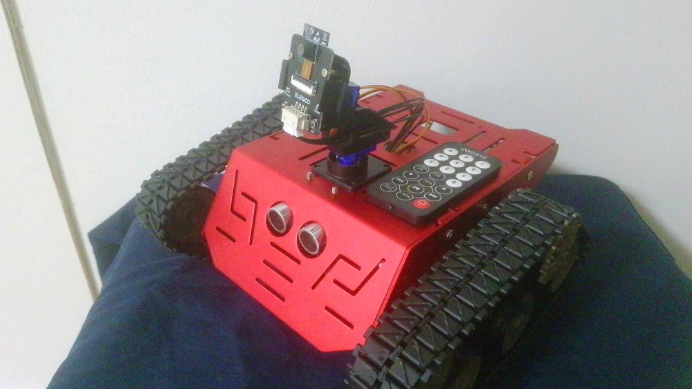 George’s assembled red ELEGOO Conqueror tank on a blue workbench surface, with tracks, camera, ultrasonic sensor, and infrared remote on its chassis.