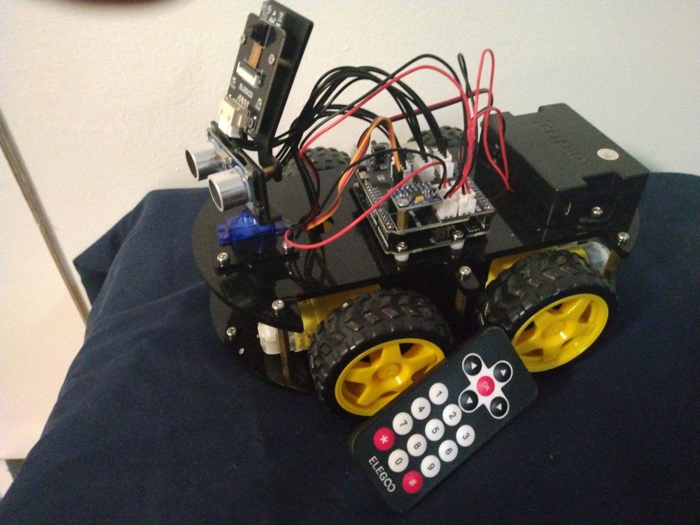 George’s assembled ELEGOO Smart Robot Car V4 on a blue workbench surface, with four yellow wheels, mounted camera and ultrasonic sensor, electronics, battery enclosure, and infrared remote.