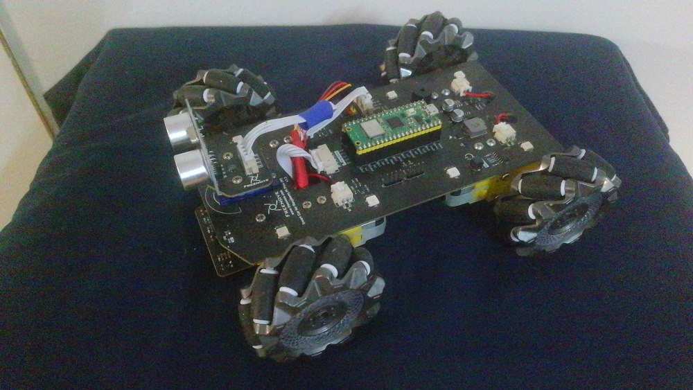 George’s assembled Freenove mecanum car on a blue workbench surface, with four roller wheels, a mounted Pico-style controller, ultrasonic sensor, display board, and wiring.